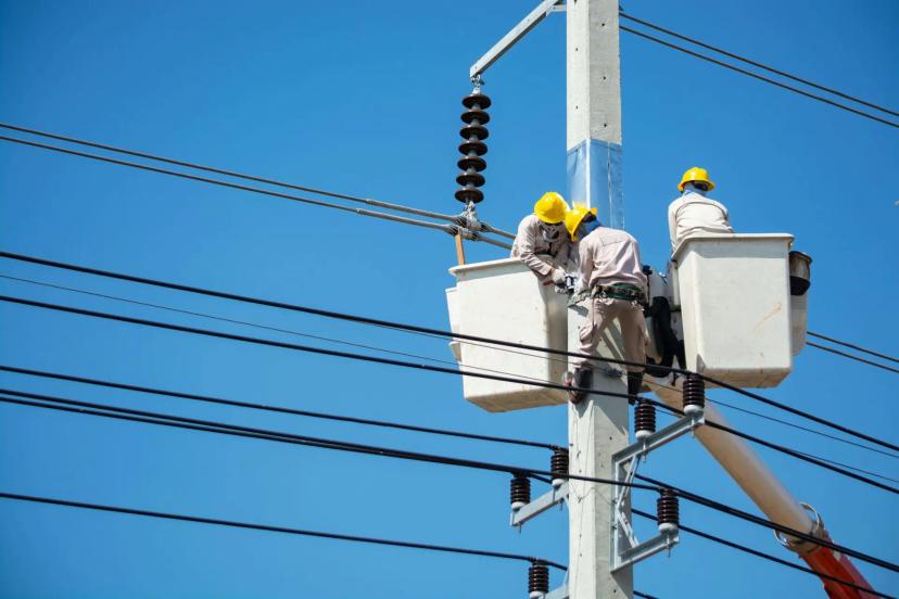 Competências digitais na manutenção de infraestrutura elétrica e redes Três eletricistas de capacete amarelo trabalhando em cestos aéreos no topo de um poste de energia sob céu azul limpo.