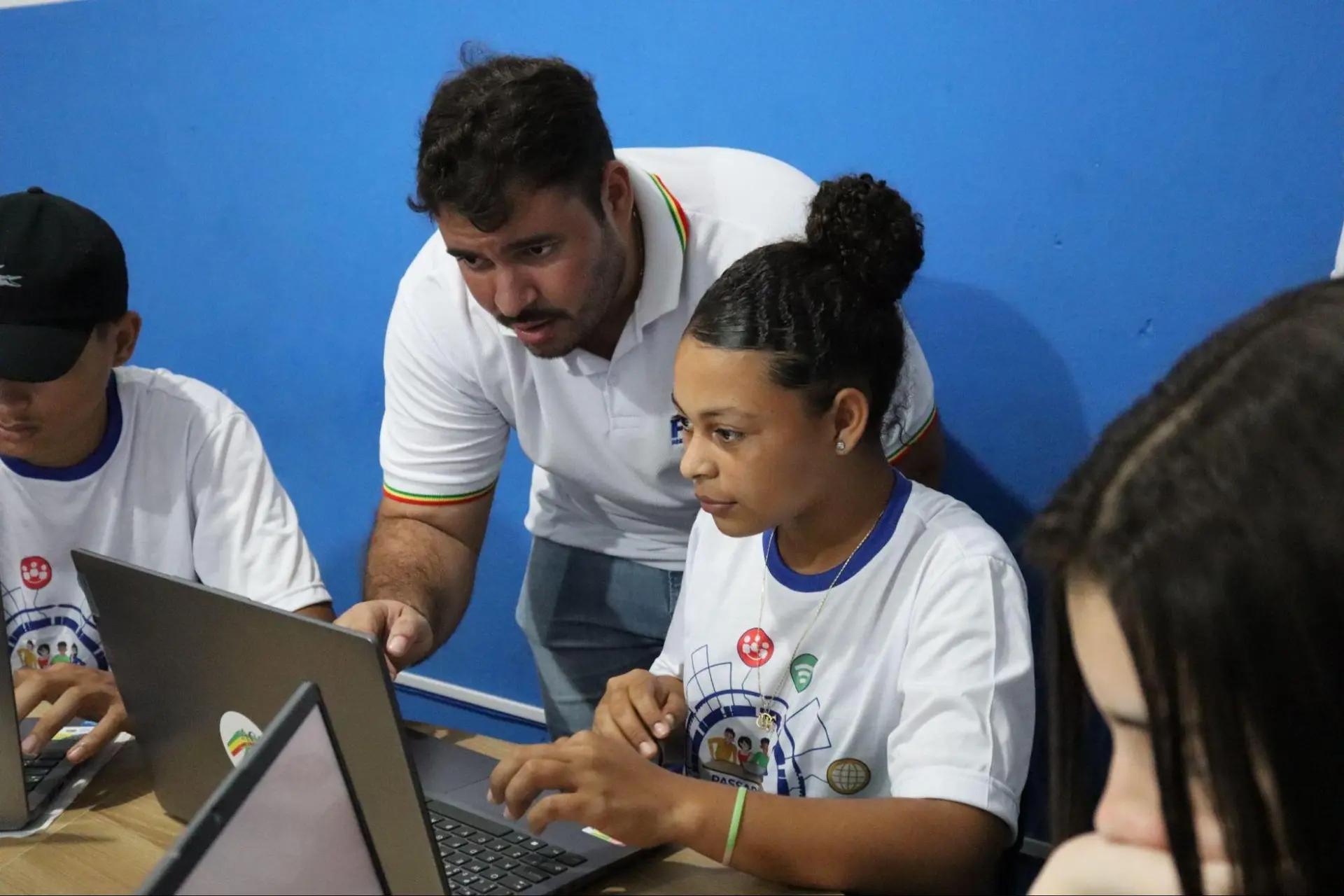 Parcerias estratégicas na educação digital entre institutos e alunos Professor instrui jovem estudante negra que utiliza laptop em sala de aula; ambos concentrados na tela em ambiente educacional.