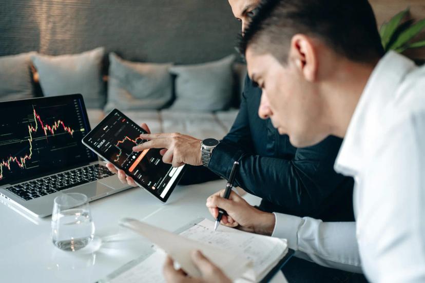IA e ML na personalização de serviços financeiros e análise de dados Dois homens analisam gráficos financeiros em um tablet e notebook, enquanto um deles faz anotações em papel sobre a mesa.