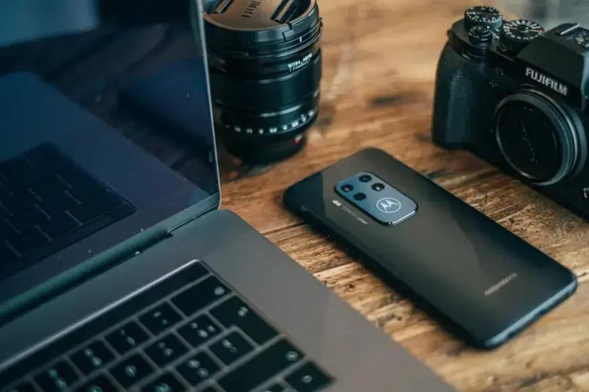 Mobile Security – Partnership between Motorola and CESAR Black Motorola smartphone on a wooden table next to an open laptop, a camera lens, and a professional camera