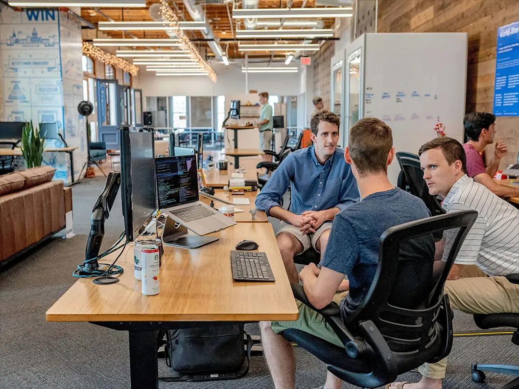 Startups e grandes empresas em parcerias estratégicas de inovação Três homens conversam em escritório moderno com portáteis, exemplificando parcerias estratégicas e colaboração profissional.