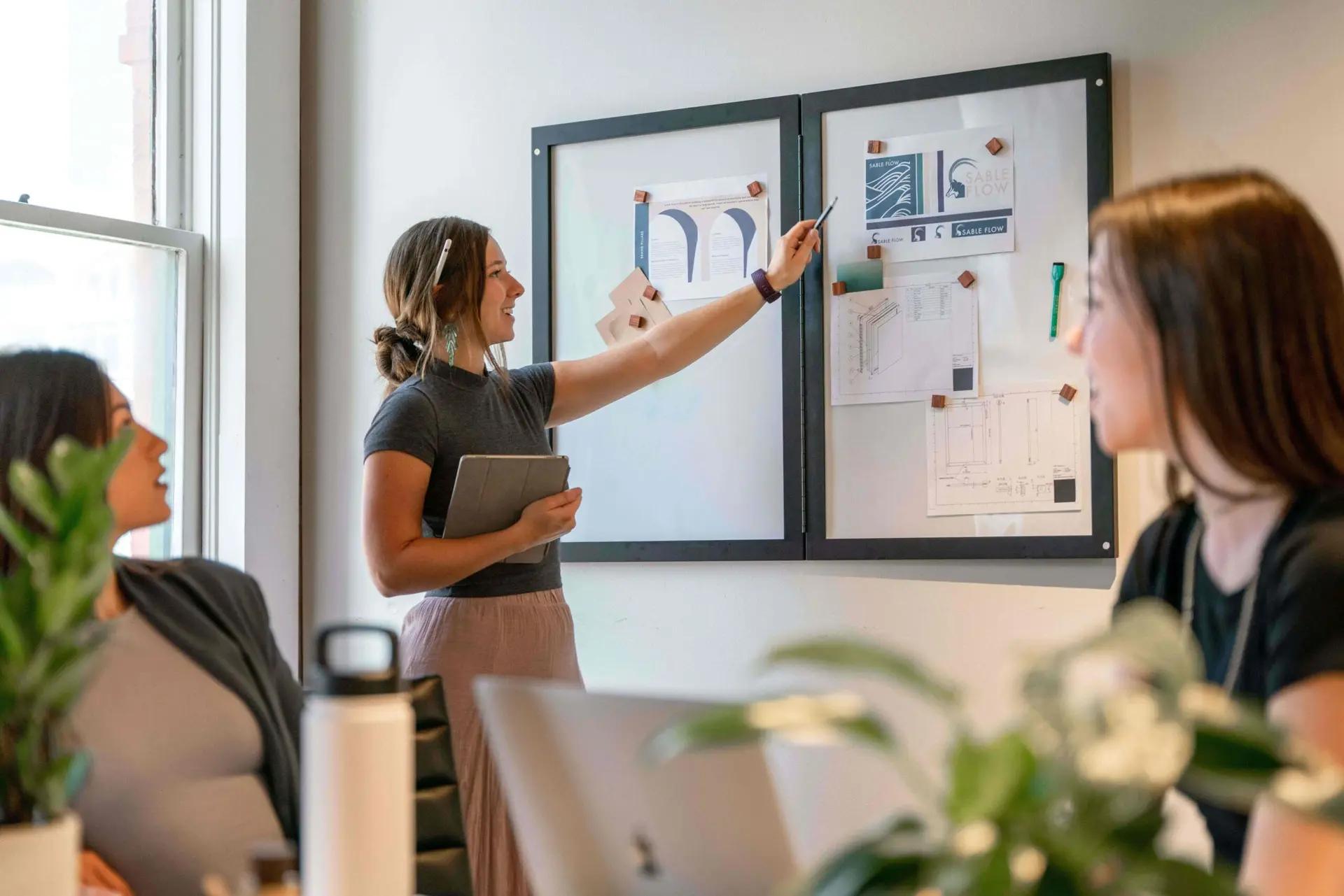 Ferramentas essenciais para promover a inovação em startups Mulher aponta para quadro branco com referências visuais durante reunião com outras duas pessoas em escritório iluminado.