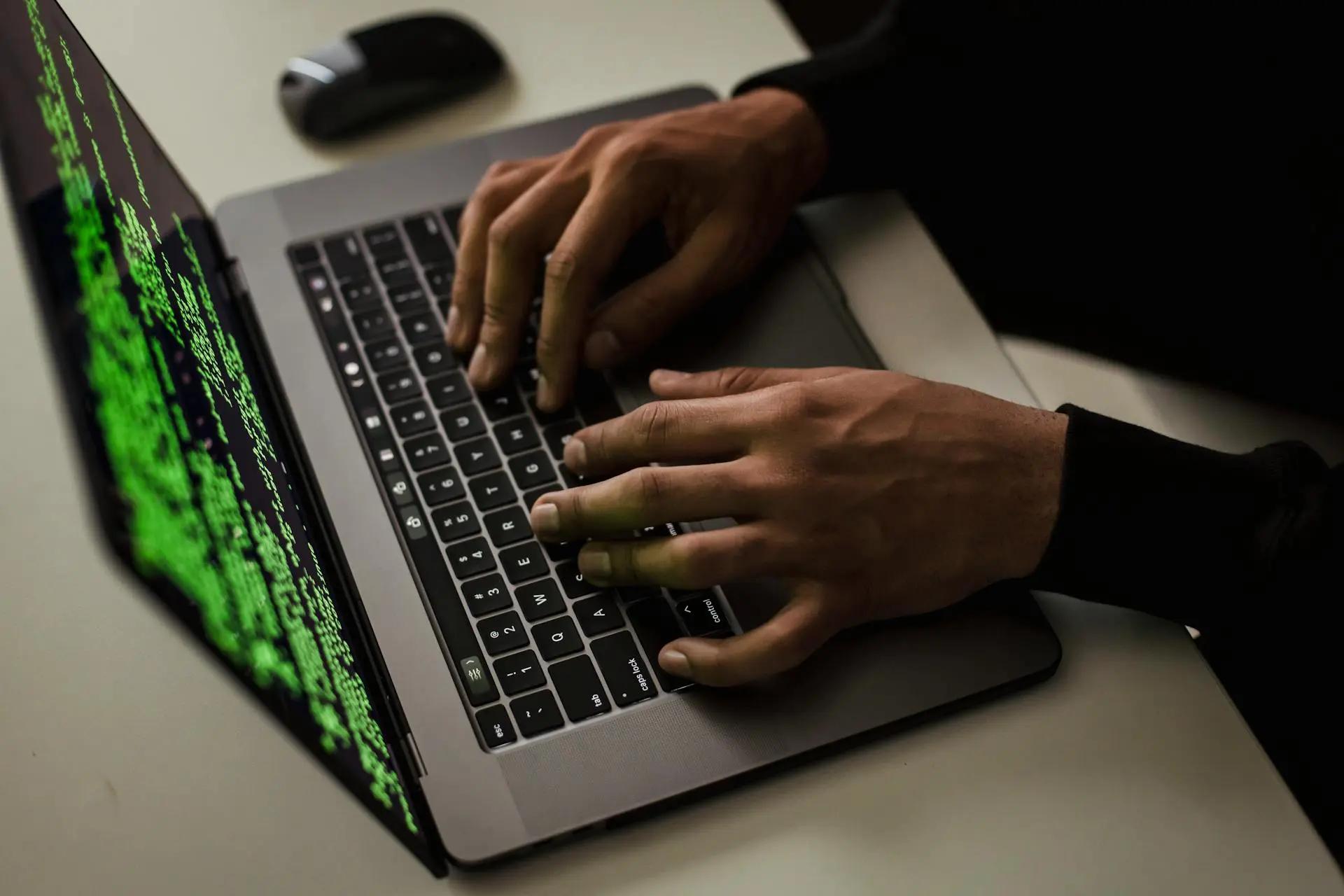Cibersegurança em meios de pagamento – desafios e soluções atuais Duas mãos digitam em um laptop cinza com linhas de código verdes na tela sobre uma mesa branca com mouse ao fundo.