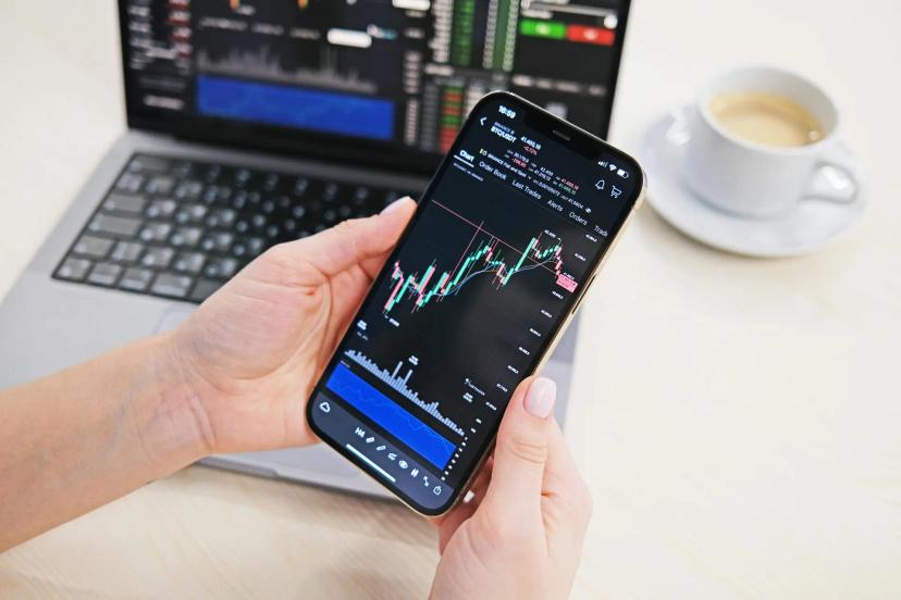 Moedas digitais e os desafios da criptografia pós-quântica Mãos segurando smartphone com gráfico de velas financeiras. Ao fundo, um laptop com dados de mercado e uma xícara de café.