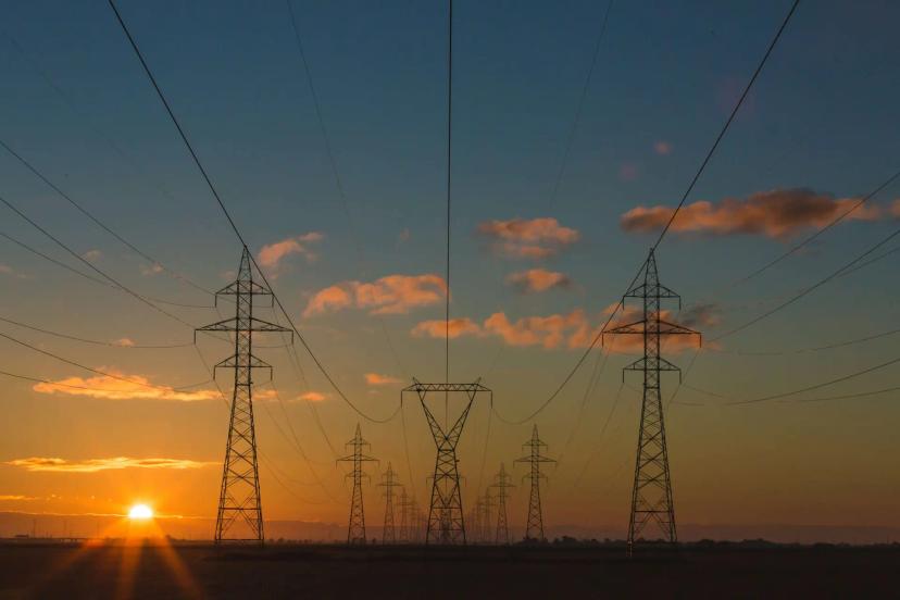 Cibersegurança em infraestruturas energéticas e proteção de redes Torres de transmissão de energia elétrica em silhueta contra um pôr do sol alaranjado com céu azul e nuvens esparsas.
