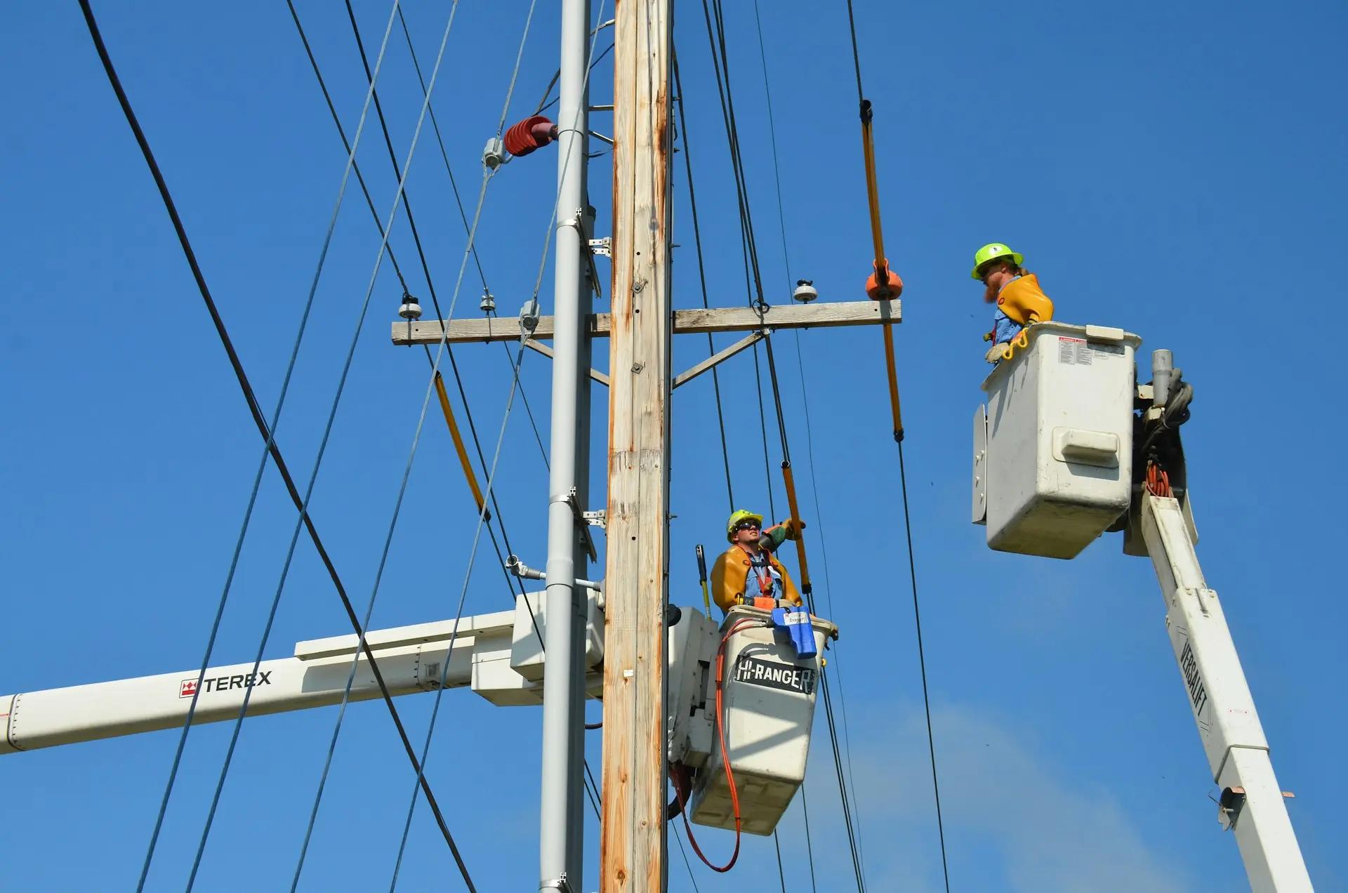 Redes inteligentes impulsionam energia verde Dois eletricistas em cestos aéreos de braços mecânicos realizam manutenção em fiação de postes elétricos sob céu azul.