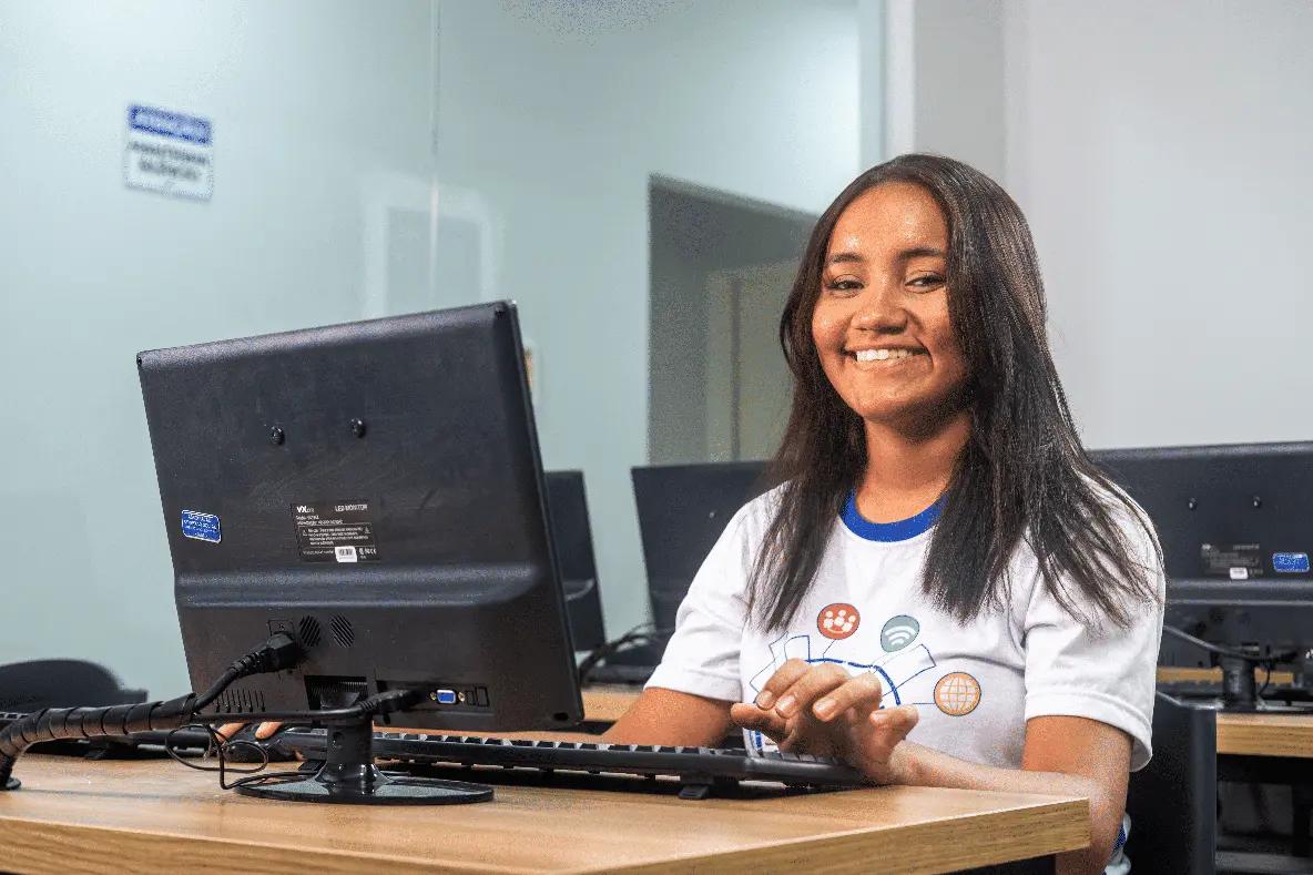 Educação inovadora impulsiona profissões do futuro e inclusão Jovem sorridente sentada em laboratório de informática com as mãos no teclado de um computador. Ao fundo, outros monitores.