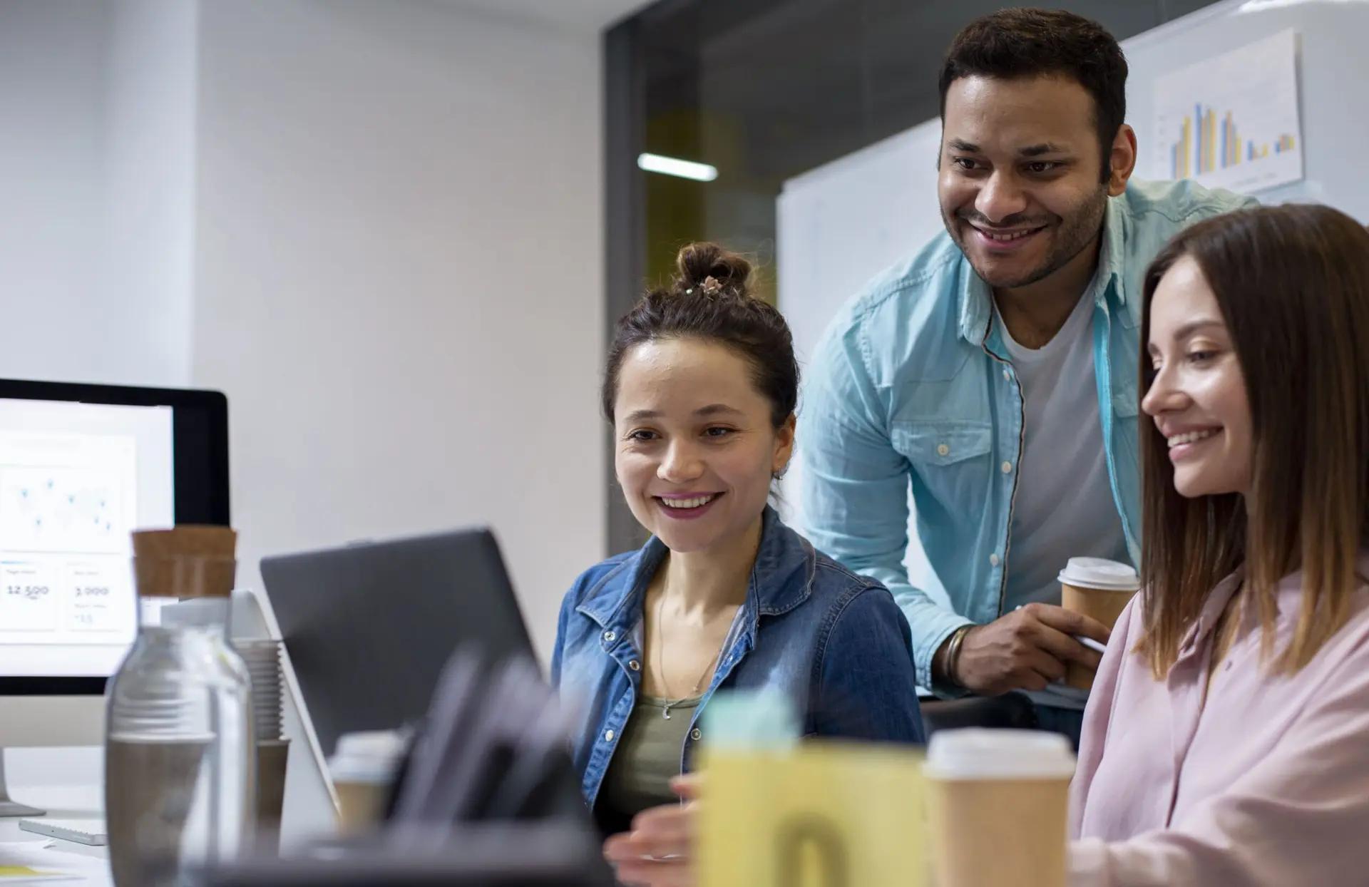 Maturidade tecnológica em startups acelerada pelo venture building Três profissionais sorridentes, duas mulheres e um homem, olham juntos para a tela de um laptop em escritório moderno.