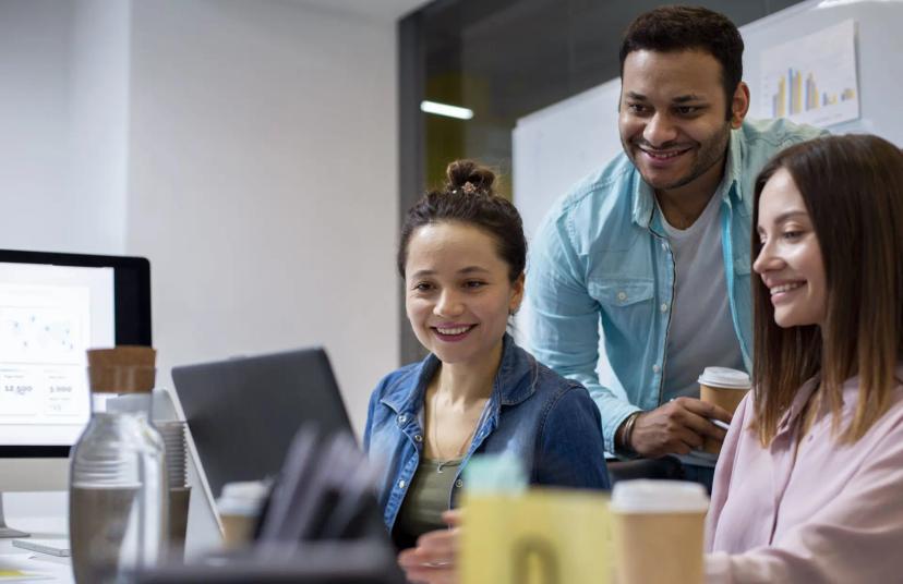 Maturidade tecnológica em startups acelerada pelo venture building Três profissionais sorridentes, duas mulheres e um homem, olham juntos para a tela de um laptop em escritório moderno.