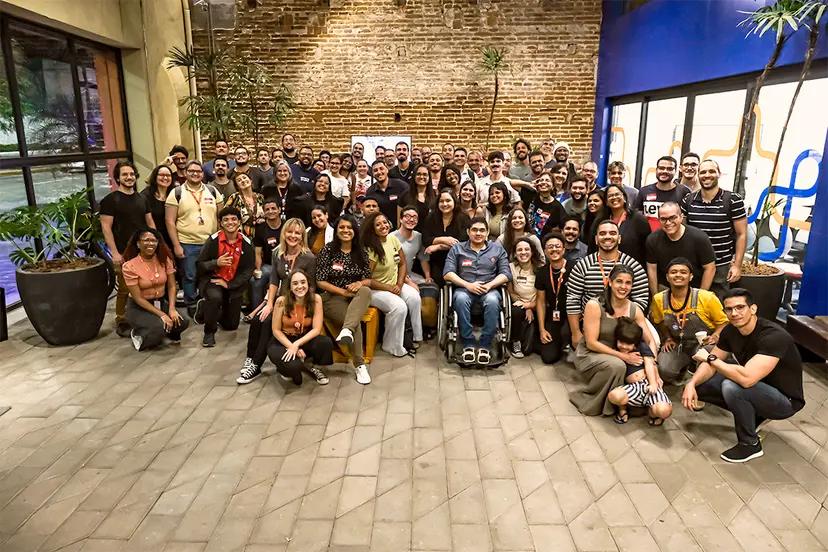 Programa FAST promove educação tecnológica e inclusão social Grupo numeroso de alunos e mentores sorrindo em um pátio com parede de tijolos.