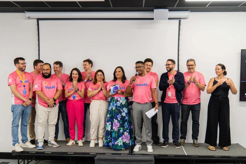 Agentes de mudança e estratégias para engajar colaboradores na inovação Grupo de doze pessoas no palco de um auditório; a maioria veste camisas rosa escrito InovAí e um homem fala ao microfone