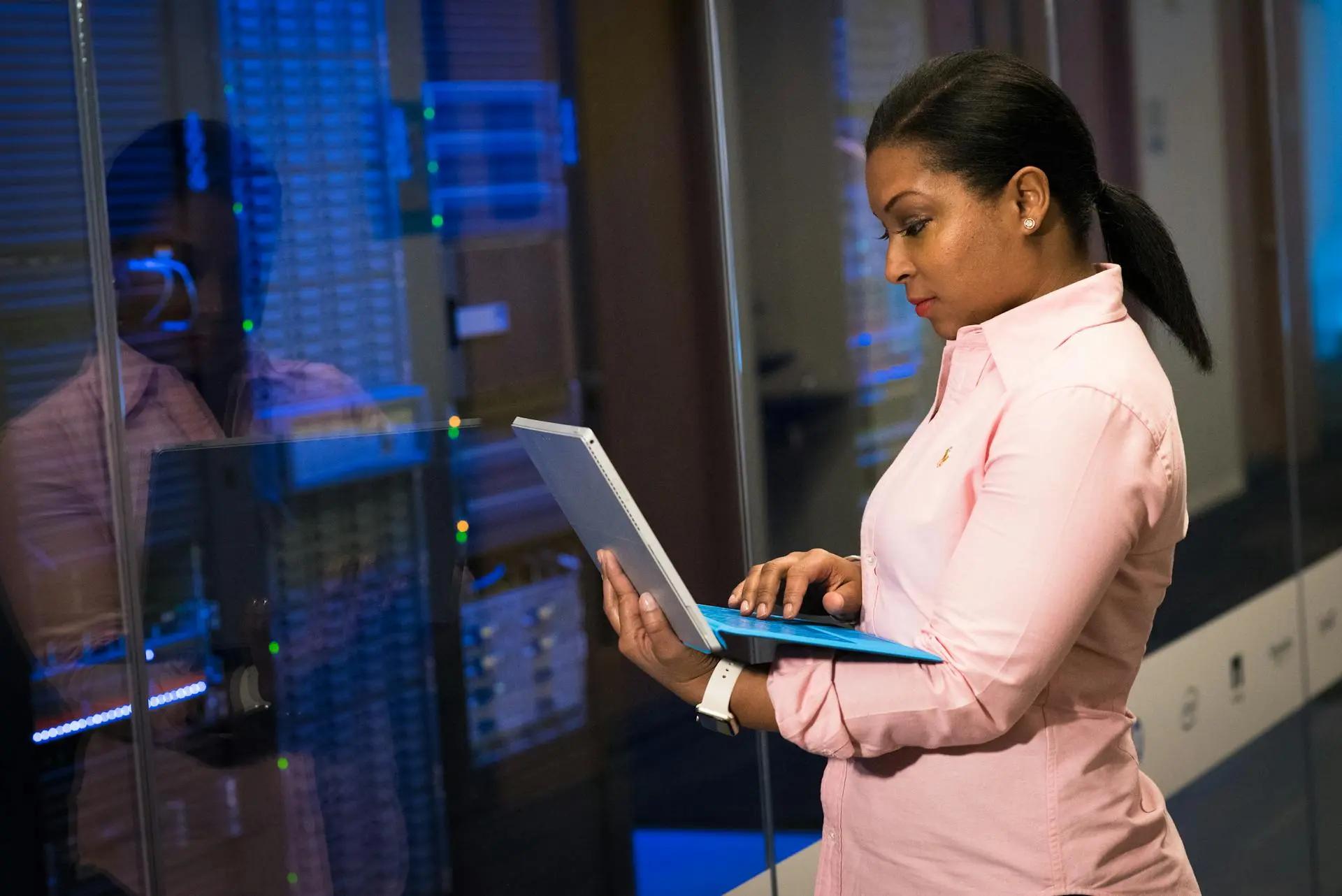 Cibersegurança na indústria 4.0 para proteção de dados e operações Mulher de camisa rosa usa um notebook em frente a racks de servidores com luzes azuis refletidas em um vidro de proteção.
