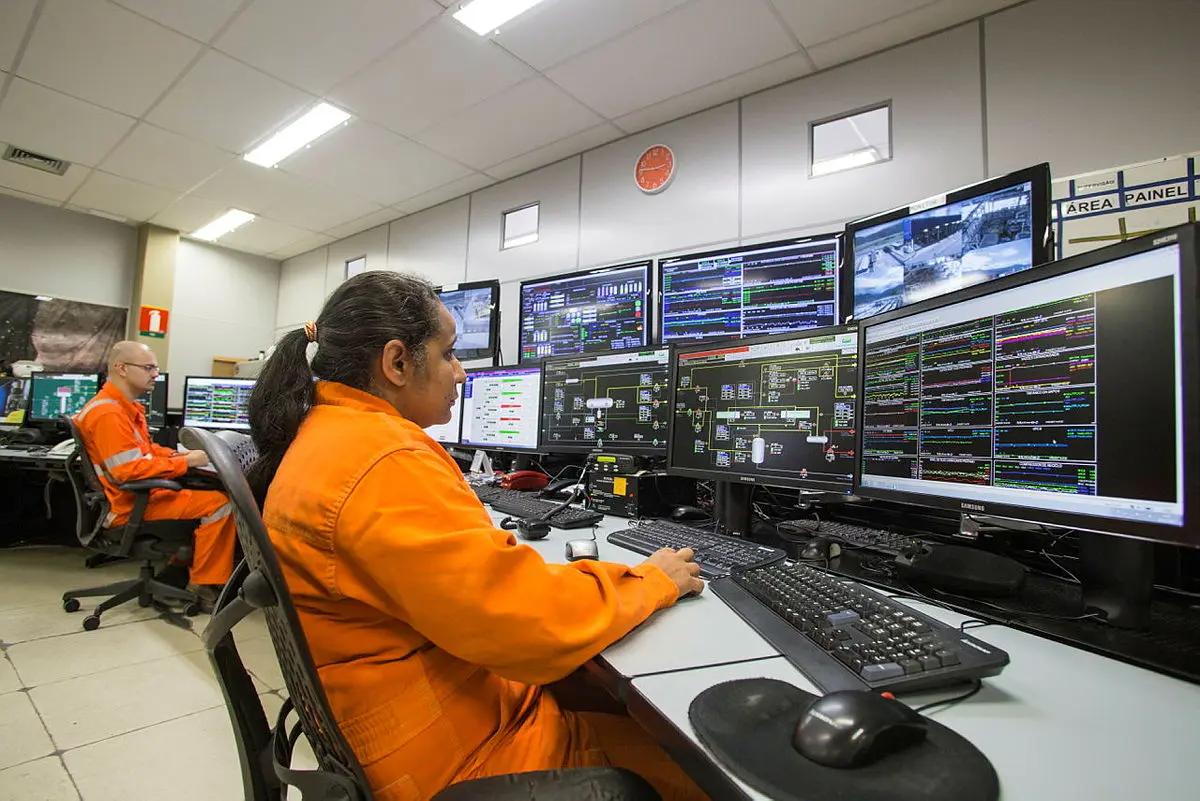 Decisões estratégicas em óleo e gás com monitoramento em tempo real Mulher e homem em macacão laranja operam computadores com múltiplos ecrãs de gráficos e mapas num centro de controlo de energia.