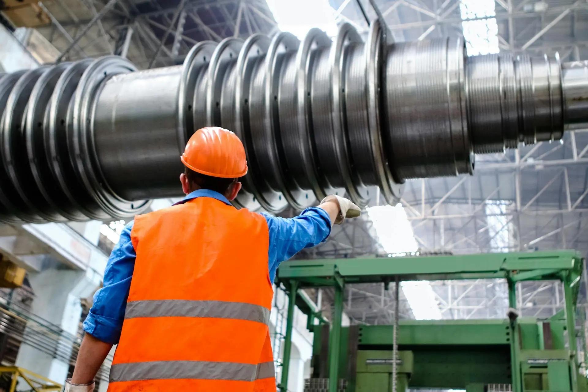 Indústria 4.0 e sustentabilidade otimizando a produção industrial Trabalhador de colete laranja e capacete aponta para uma grande turbina metálica em uma fábrica de alta tecnologia.
