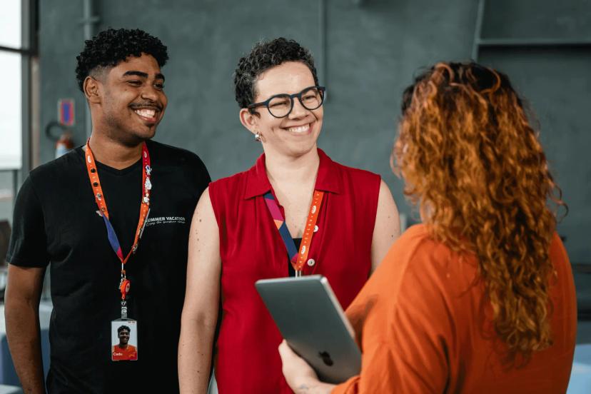Inovação corporativa com colaboração e estratégia para competitividade Três pessoas sorrindo em uma conversa de trabalho; um homem e uma mulher de crachá ouvem uma colega que segura um tablet.