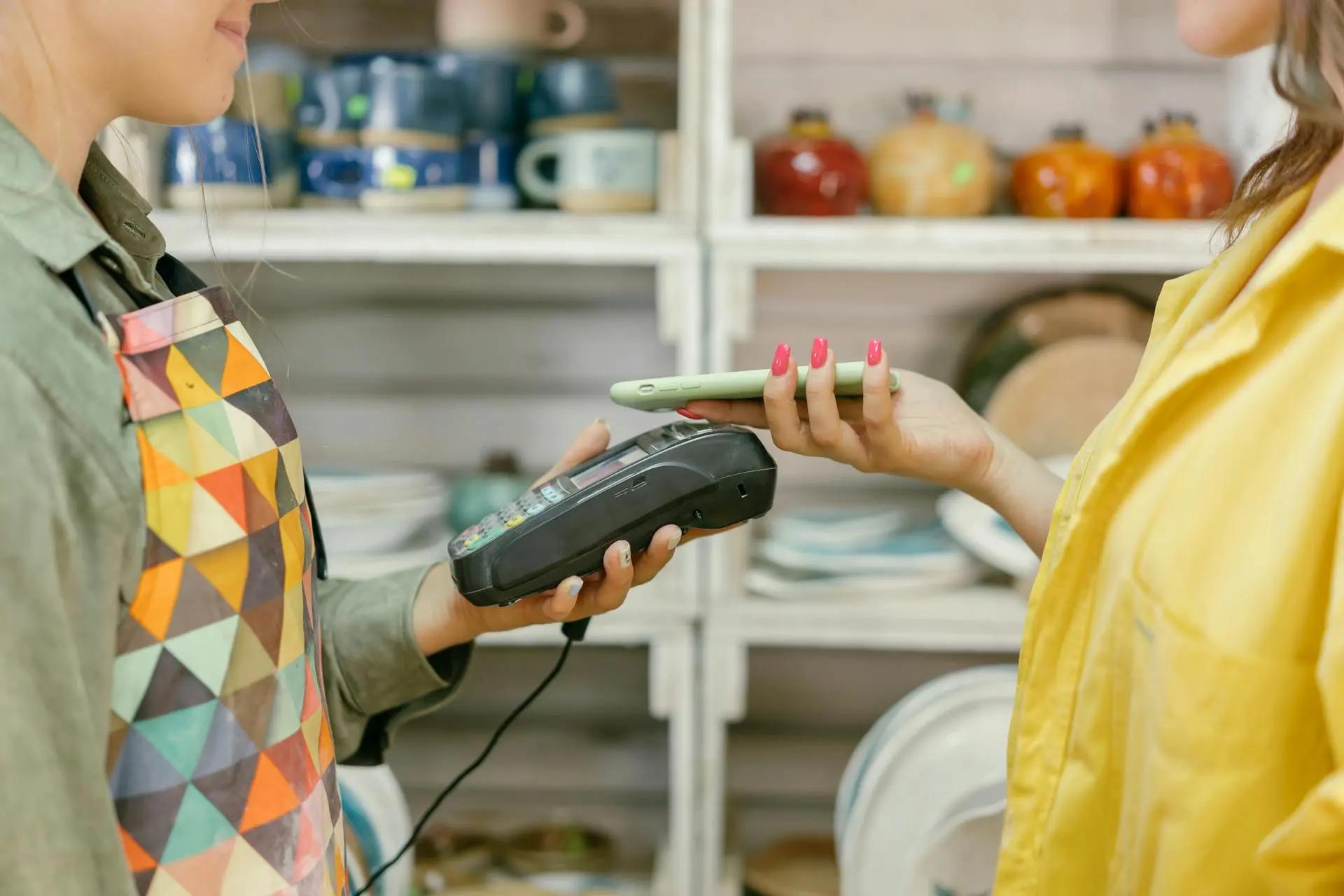 Pagamentos sem contato e o futuro das transações cashless no varejo Cliente aproxima celular de máquina de cartão segurada por atendente em loja de cerâmicas; prateleiras com canecas ao fundo.