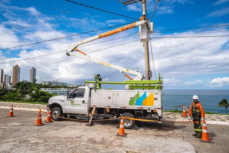 Redução de custos no setor de energia com inovação em operações Caminhão de manutenção com cesto elevatório operado por eletricista em poste; área isolada por cones junto à orla marítima.