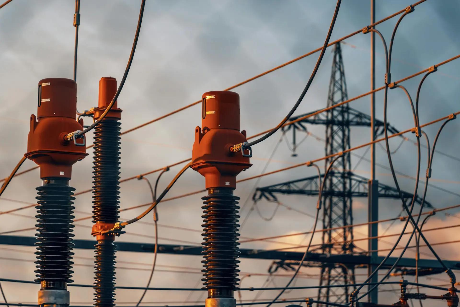 Tecnologias para eficiência energética em subestações de energia Isoladores de cerâmica e equipamentos de subestação com cabos de alta tensão e torre metálica ao fundo sob céu nublado.