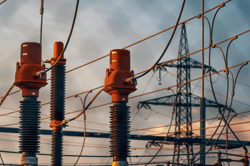 Tecnologias para eficiência energética em subestações de energia Isoladores de cerâmica e equipamentos de subestação com cabos de alta tensão e torre metálica ao fundo sob céu nublado.