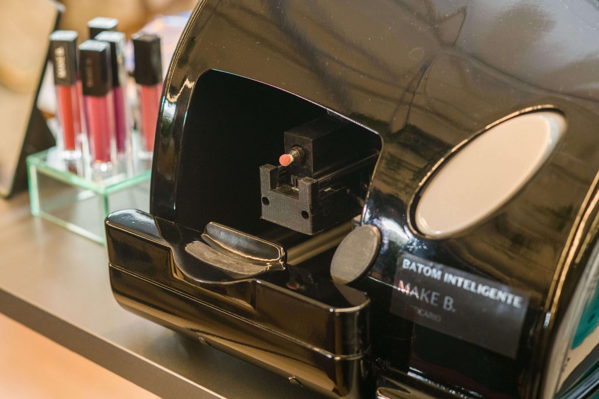 batom-inteligente-cesar-boticari Máquina de batom inteligente preta com aplicador visível, próxima a amostras de batons em um suporte.