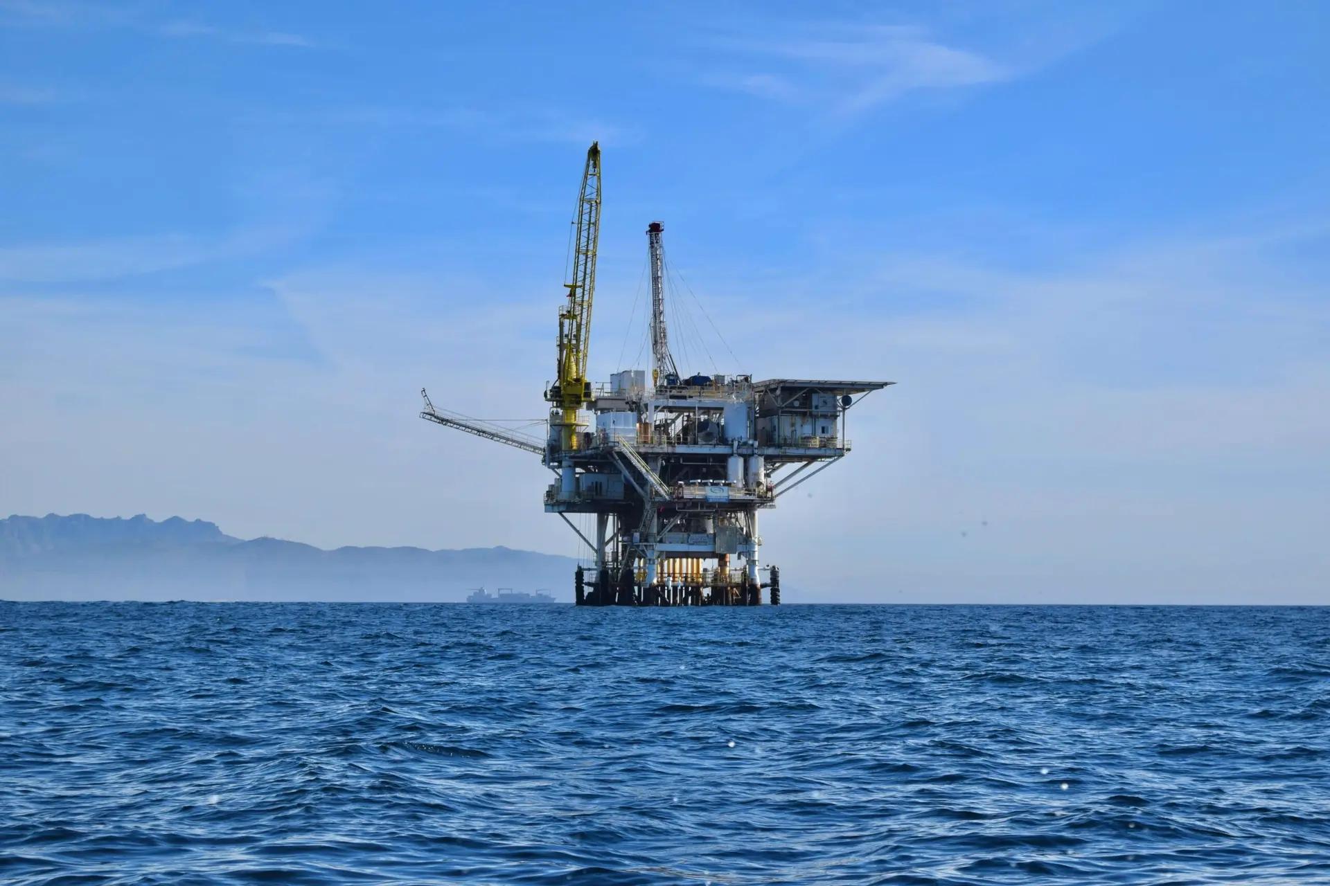 Digital Twins e a otimização de operações offshore em óleo e gás Plataforma de petróleo offshore com guindaste amarelo no centro do oceano sob céu azul; montanhas ao fundo no horizonte.