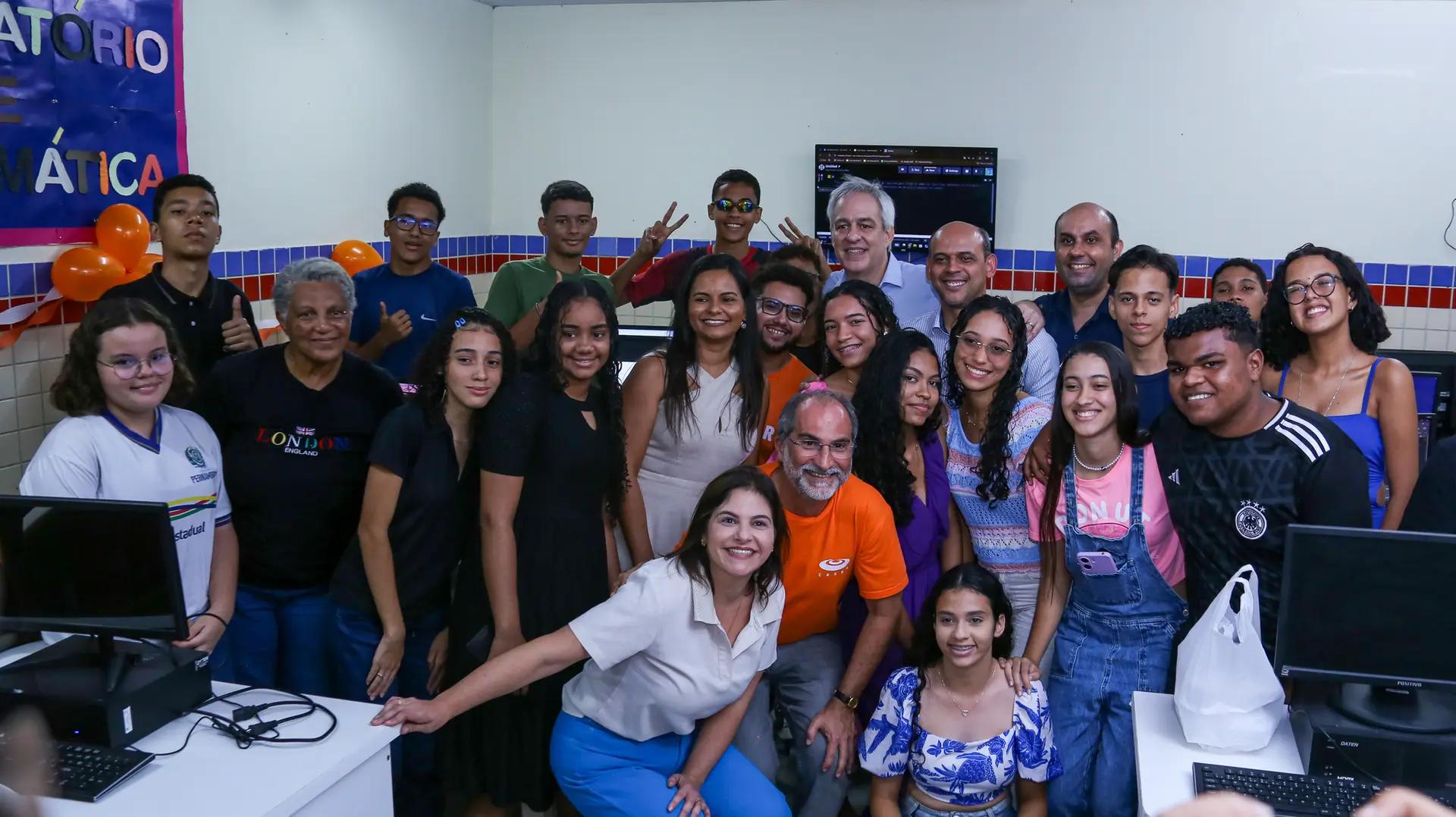 Educação digital e inclusão através de parcerias público-privadas Grupo grande de estudantes e professores sorrindo em laboratório de informática com computadores e balões decorativos.