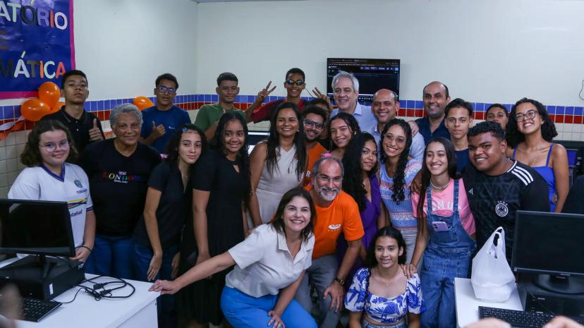 Educação digital e inclusão através de parcerias público-privadas Grupo grande de estudantes e professores sorrindo em laboratório de informática com computadores e balões decorativos.