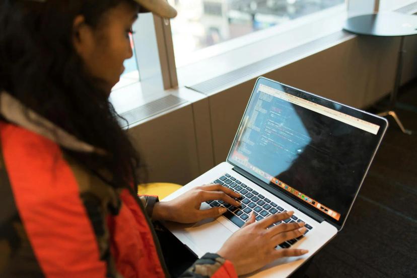 Gêmeos digitais e tecnologias da indústria 4.0 na prática Mulher com jaqueta vermelha digita códigos de programação em um notebook prata; tela exibe linhas de comando em ambiente iluminado.