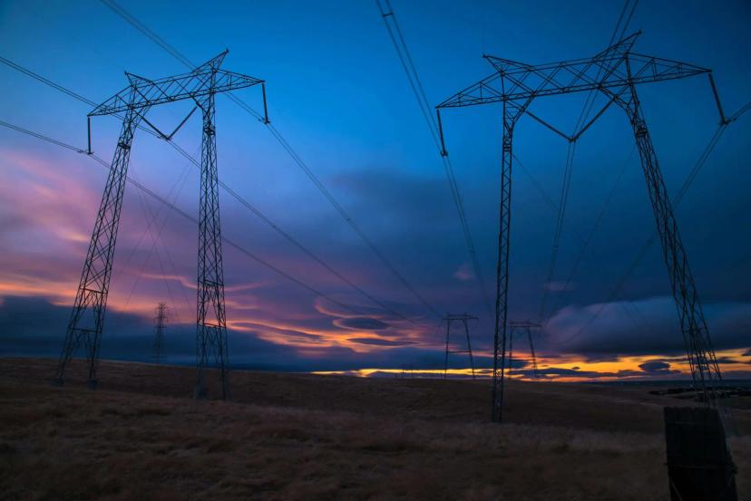 Gestão de energia elétrica e estratégias para mitigar falhas no setor Duas grandes torres de transmissão de energia em um campo aberto sob céu ao entardecer com tons de azul, rosa e laranja.