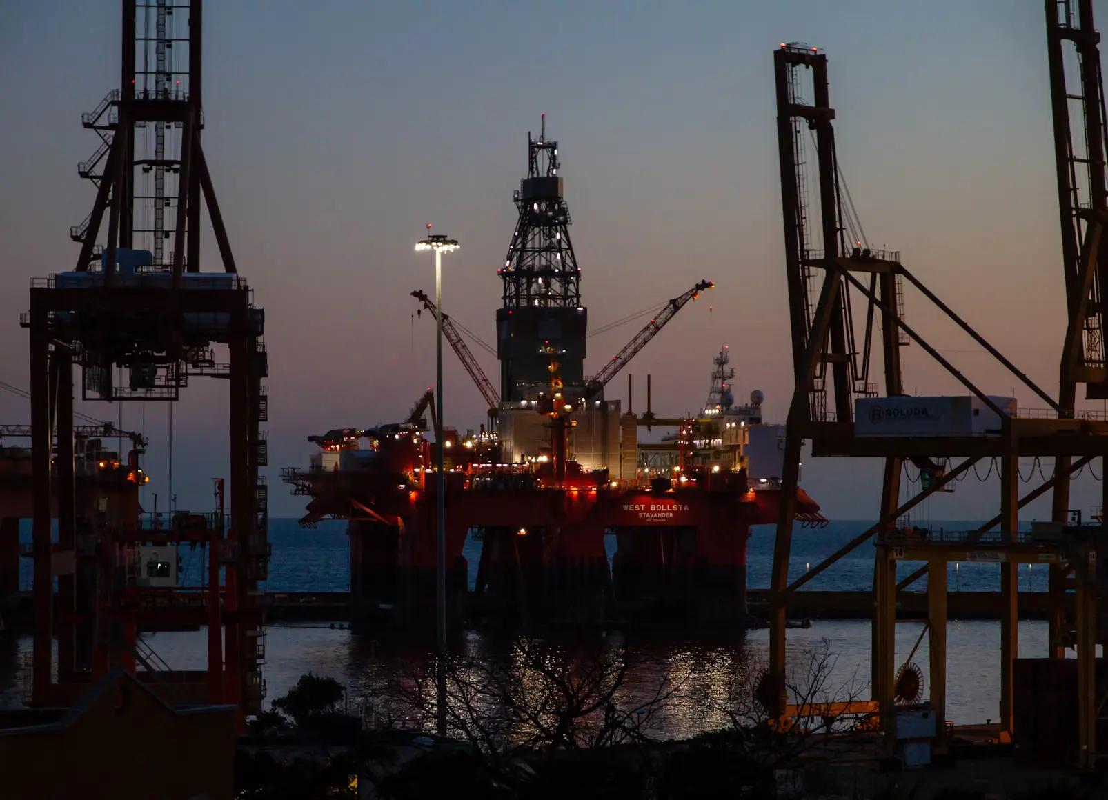 IA para ativos críticos em indústrias de óleo e gás e manutenção Plataforma de petróleo iluminada ao anoitecer, vista entre guindastes portuários em um terminal marítimo sob céu crepuscular