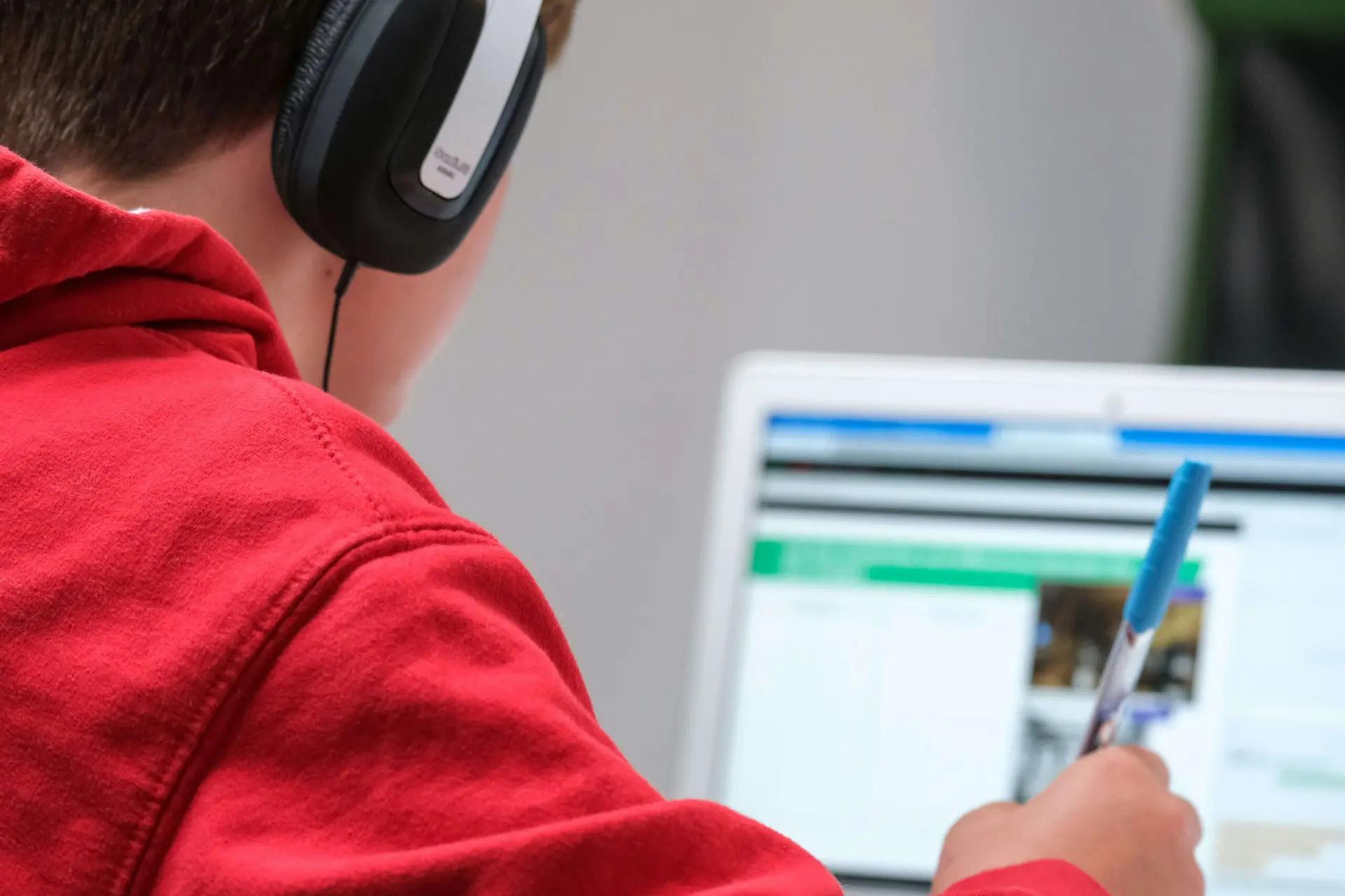 Inclusão produtiva e o papel da educação na geração de oportunidades Jovem de casaco vermelho e fones de ouvido estuda em frente ao computador enquanto faz anotações com uma caneta azul.