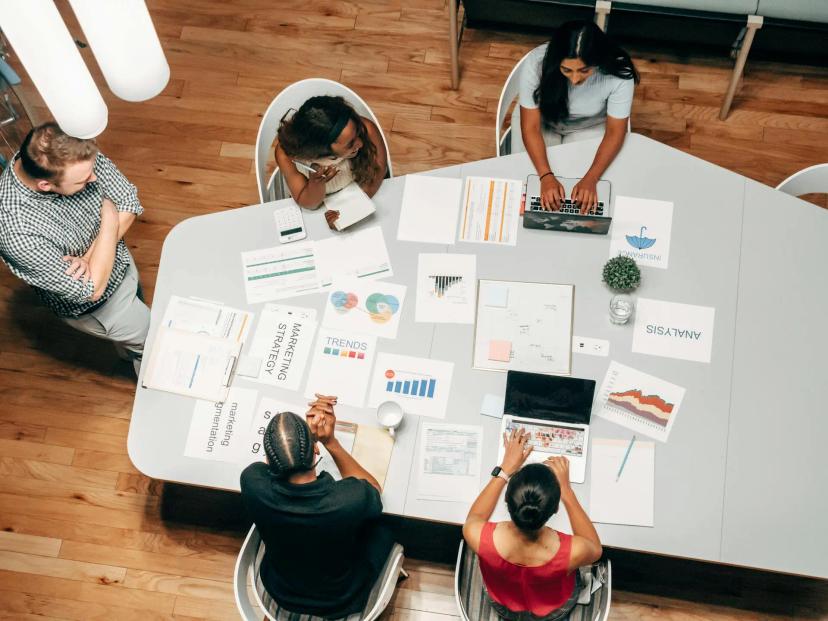 Inovação corporativa e os riscos de não investir em novas estratégias Cinco pessoas em reunião ao redor de mesa com laptops, gráficos de tendências, post-its e papéis de estratégia de marketing.
