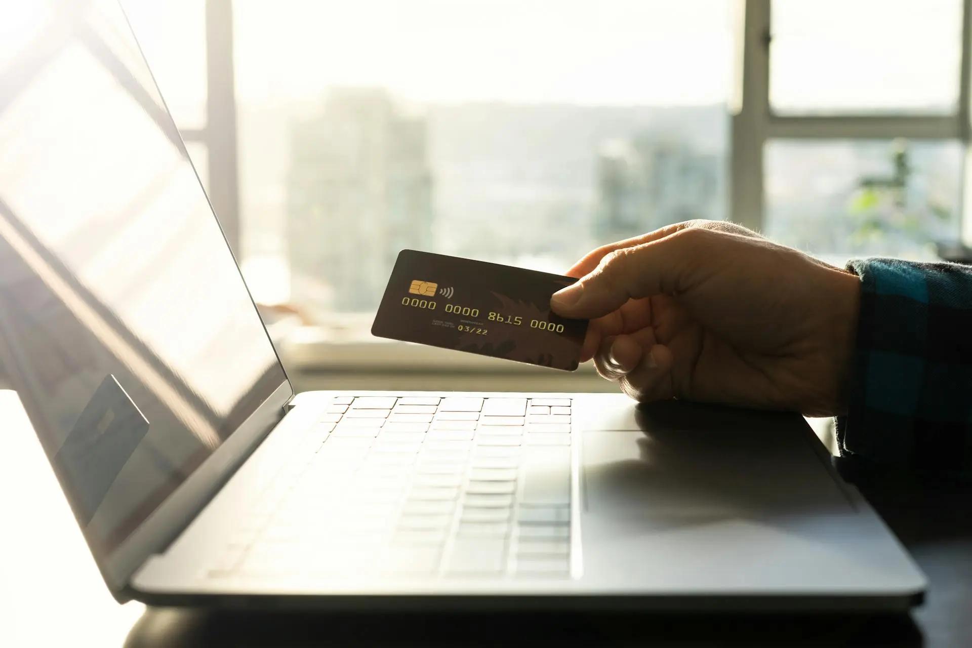 Cibersegurança financeira e os riscos invisíveis em transações digitais Mão segurando cartão de crédito preto em frente a um notebook aberto, próximo a uma janela iluminada pelo sol