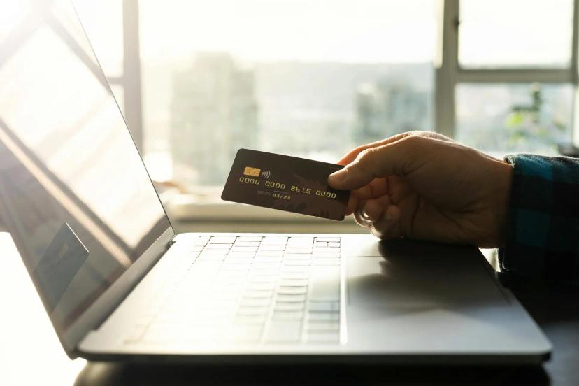 Cibersegurança financeira e os riscos invisíveis em transações digitais Mão segurando cartão de crédito preto em frente a um notebook aberto, próximo a uma janela iluminada pelo sol