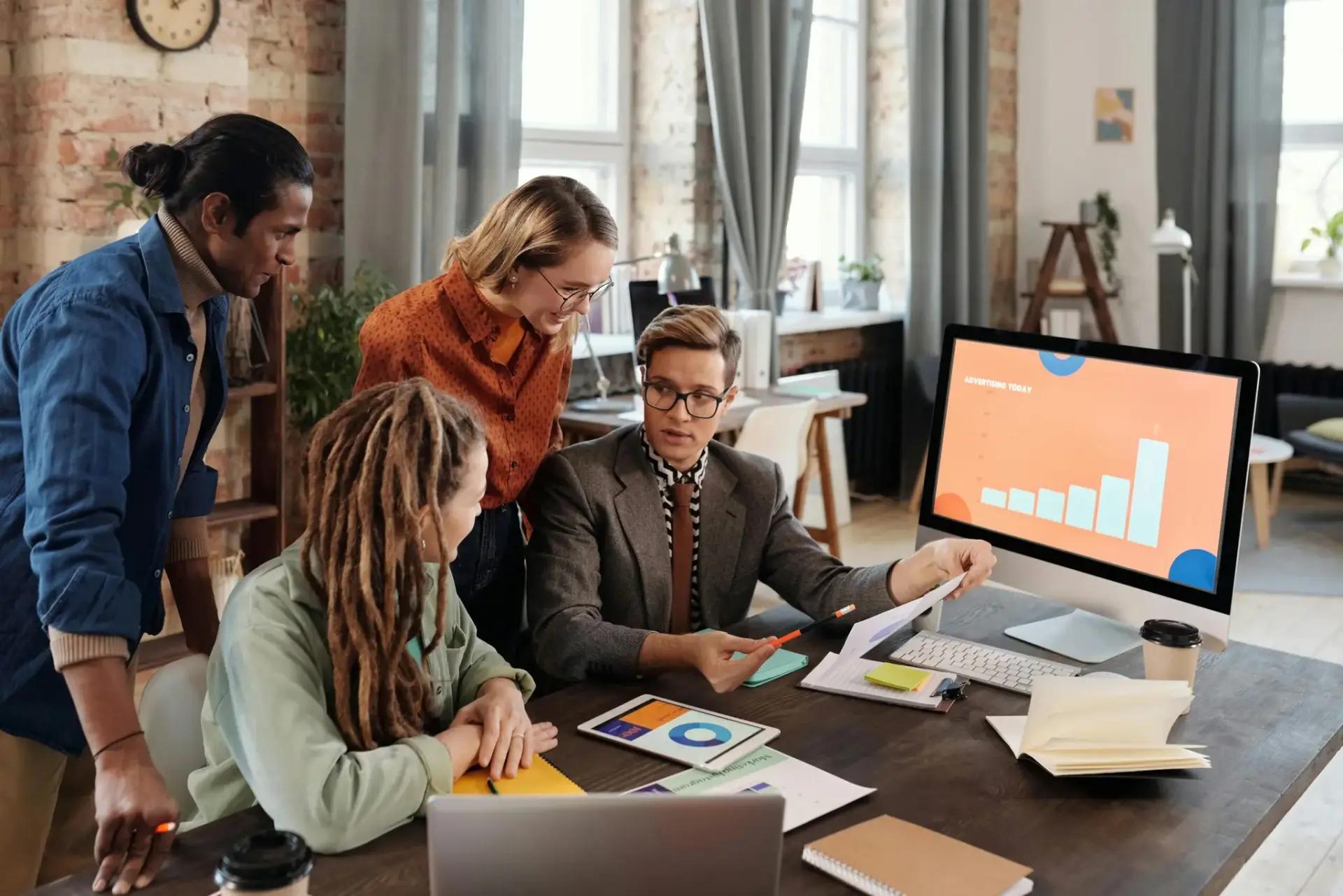 Equipe de P&D e a importância estratégica de designers no comando Quatro profissionais ao redor de uma mesa analisam gráficos em um monitor e tablet em escritório moderno e colaborativo.
