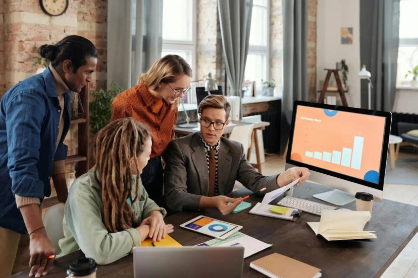 Equipe de P&D e a importância estratégica de designers no comando Quatro profissionais ao redor de uma mesa analisam gráficos em um monitor e tablet em escritório moderno e colaborativo.