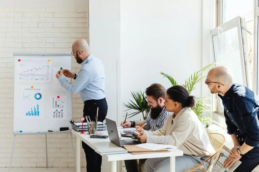 Rodadas de investimento para inovação em startups e análise financeira Quatro pessoas em reunião de planejamento. Homem em pé aponta para quadro com gráficos financeiros enquanto equipe faz anotações.
