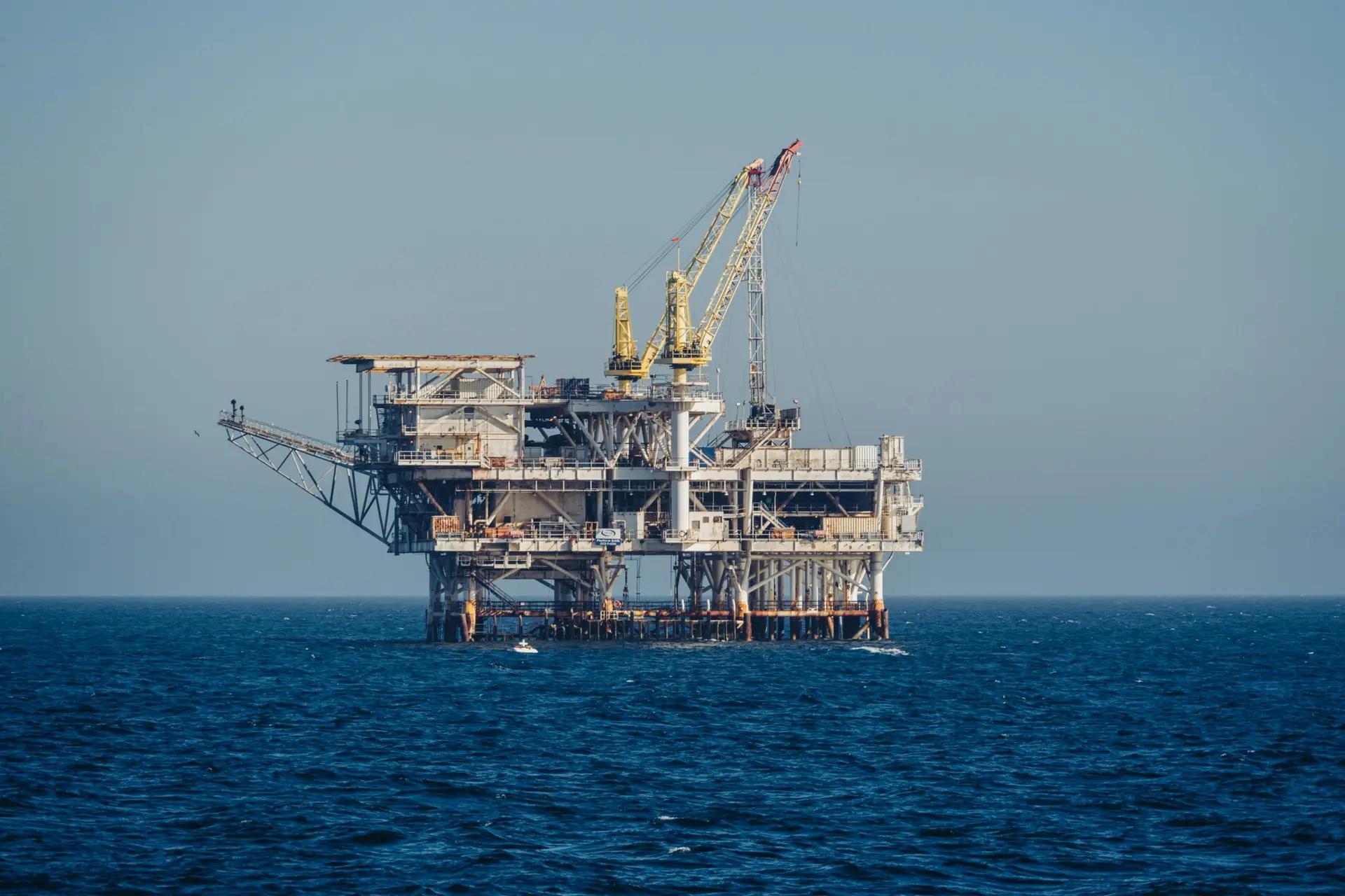 Dados e tecnologia otimizando a inteligência operacional em óleo e gás Plataforma de petróleo offshore com guindastes amarelos em operação no meio do oceano sob um céu azul limpo e ensolarado.