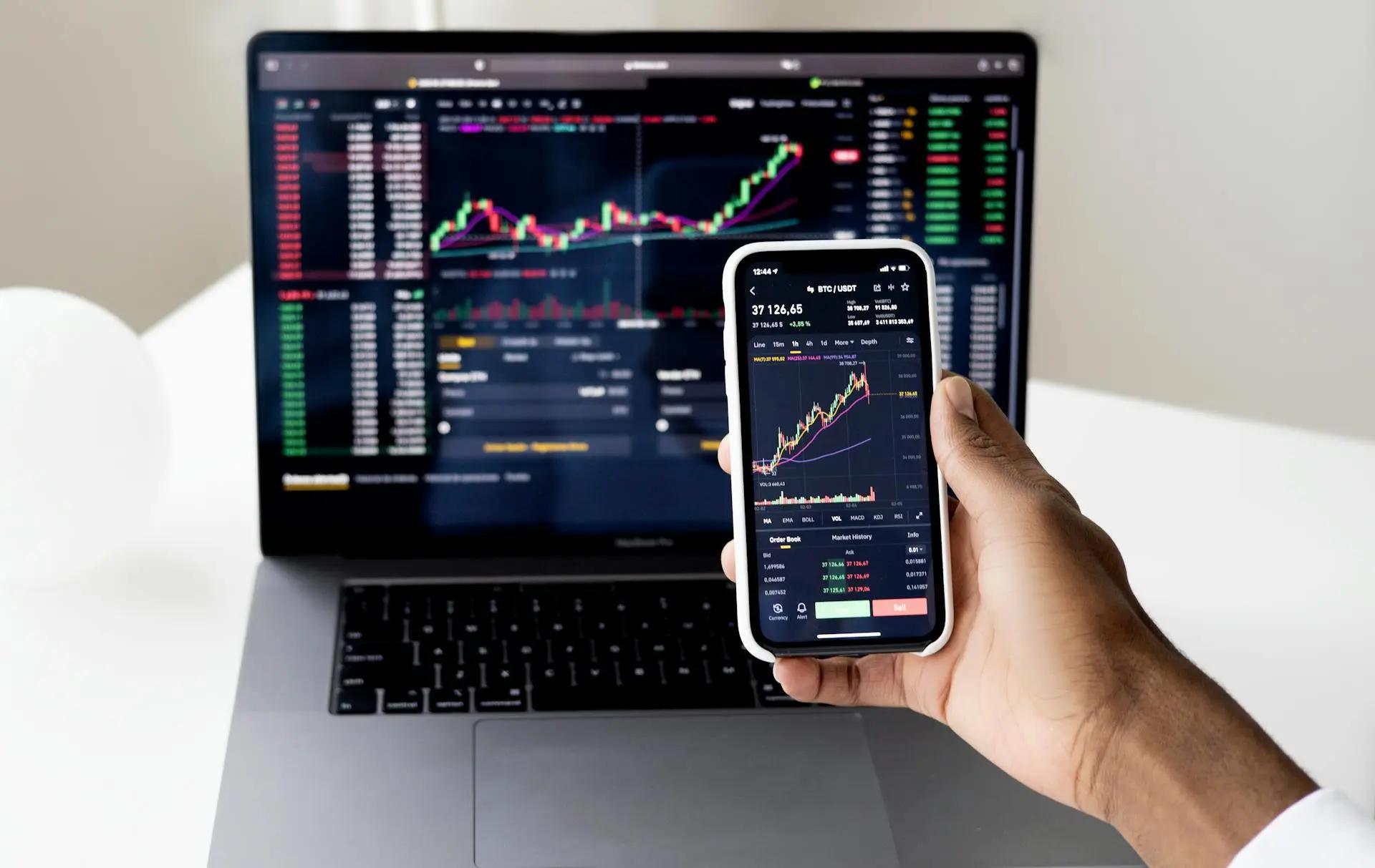 IA e personalização no setor bancário para prever desejos do cliente Mão segurando smartphone com gráficos financeiros de criptomoedas em frente a um notebook exibindo dados de mercado similares.