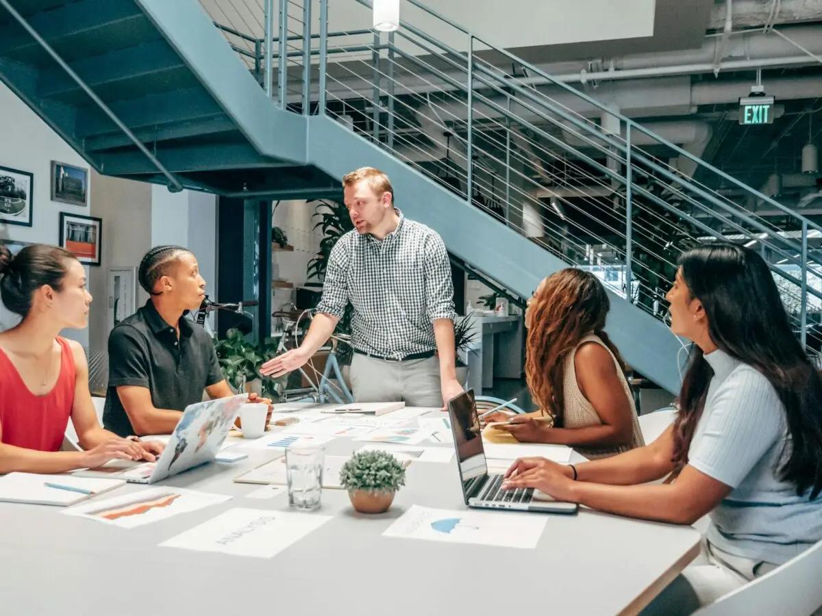 Laboratório de inovação e o fomento à cultura de experimentação Grupo de cinco pessoas em reunião colaborativa em escritório moderno. Homem em pé lidera conversa ao redor de mesa com laptops.