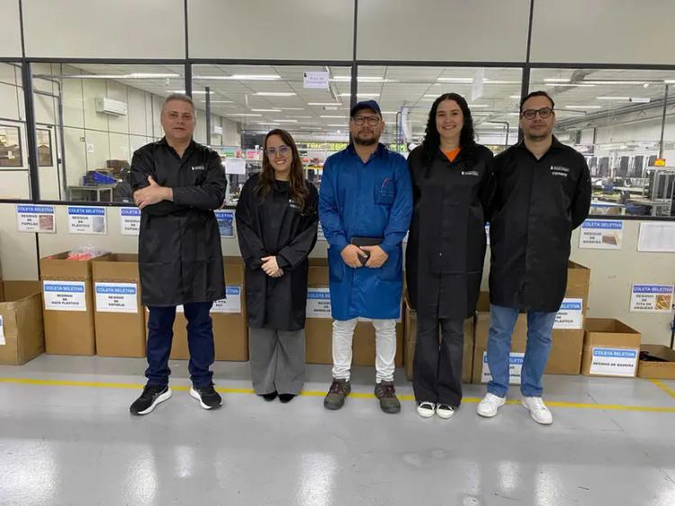 Boardtec automatiza controle de paradas para gestão em tempo real Cinco profissionais de jaleco posam alinhados em fábrica, à frente de caixas de coleta seletiva e divisória de vidro
