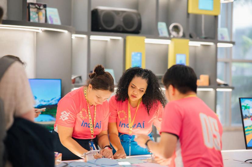 Educação corporativa – Estratégia para vantagem competitiva e inovação Duas mulheres de camiseta rosa escritas InovAí e logo do Banco do Brasil trabalham juntas em bancada em ambiente moderno.