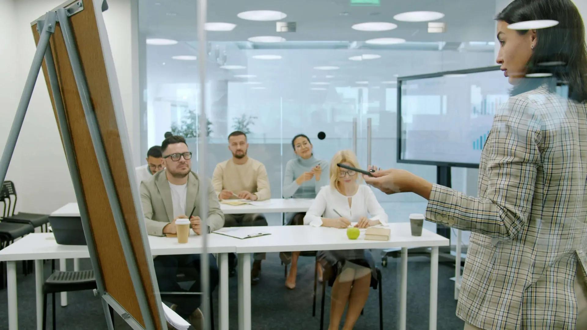 Educação corporativa com dados e IA para performance de equipes Uma mulher palestrante apresenta para cinco pessoas sentadas em uma sala moderna. Há um quadro branco à esquerda e monitor ao fundo.