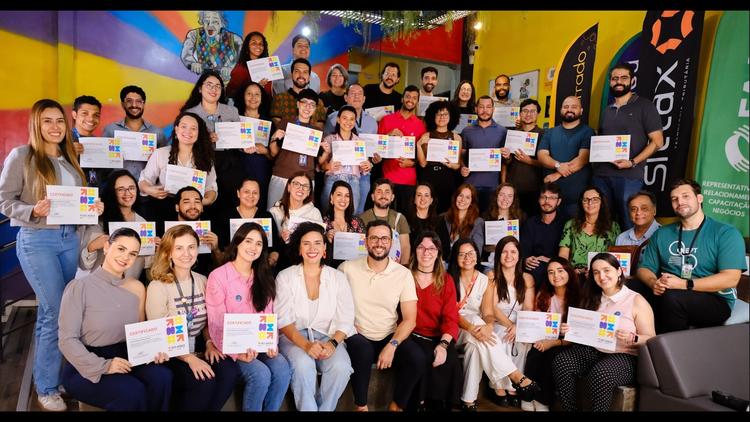 Formação em agilidade para servidores públicos Grupo de aproximadamente 40 servidores sorridentes exibe certificados coloridos em um ambiente moderno e vibrante