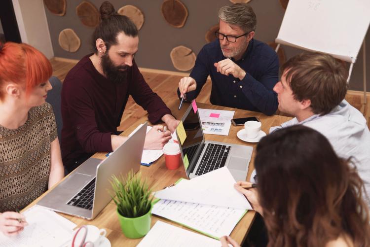 Startups e Ventures impulsionam a tecnologia no mundo dos negócios : Cinco pessoas em mesa de madeira analisam gráficos em papéis e laptops. Há post-its coloridos e plantas no escritório.