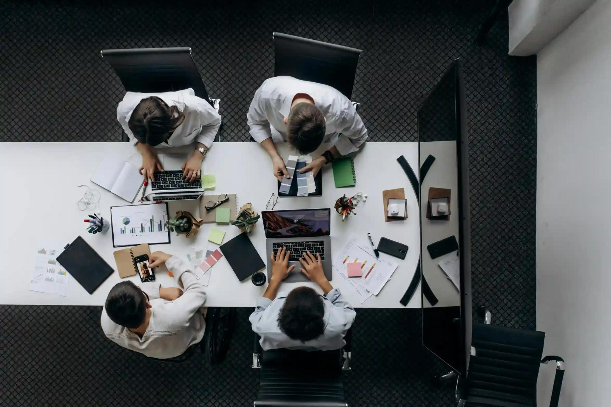Ventures e tecnologia para escalar negócios com colaboração Três profissionais em escritório colaborativo, um no computador enquanto duas colegas analisam um caderno ao fundo
