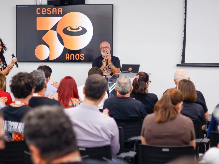 CESAR 30 anos celebrando três décadas de inovação e impacto tecnológico Silvio Meira em um púlpito diante de uma plateia sentada, ao lado de uma grande tela de TV que exibe o logotipo comemorativo "CESAR 30 ANOS"