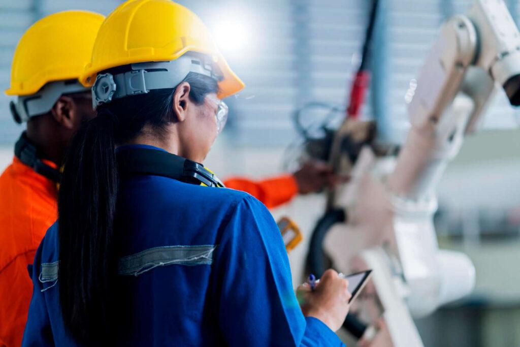 Engenheira de capacete amarelo e uniforme azul faz anotações em prancheta digital ao monitorar braço robótico industrial com colega.