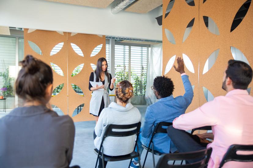 Engajamento de equipes Pessoas participando de uma atividade em grupo, com uma mulher apresentando e um participante levantando a mão em um ambiente corporativo