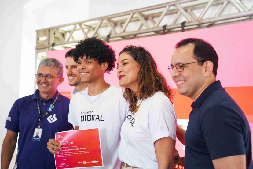 Impacto social por meio da educação digital Jovem sorridente segura certificado em cerimônia com equipe vestindo camisetas do programa de fundação digital.